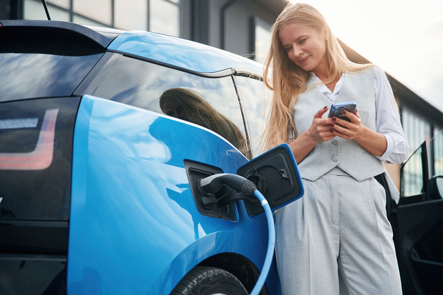 A woman stands beside a bright blue electric car while it charges, holding her smartphone and smiling. The charging cable is plugged into the car’s port near the rear wheel. She is dressed in light gray business attire and appears relaxed, possibly checking her phone as the vehicle charges in an urban setting with modern buildings reflected on the car’s surface.
